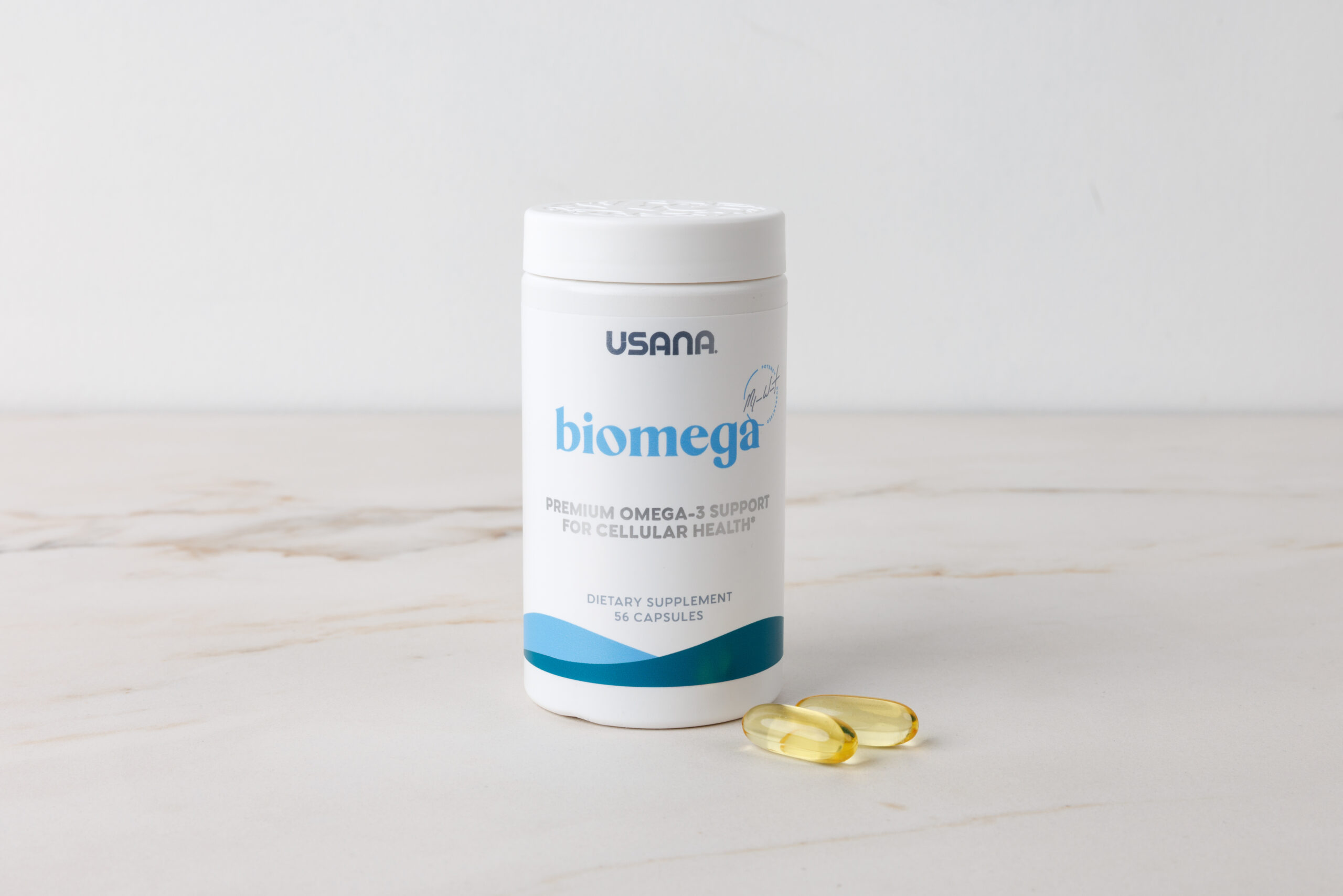 一瓶白色 USANA Biomega 產品,標籤註明優質 Omega-3 細胞健康支援,瓶身旁有兩顆透明金黃色魚油膠囊,背景為簡約乾淨的室內自然光風格。